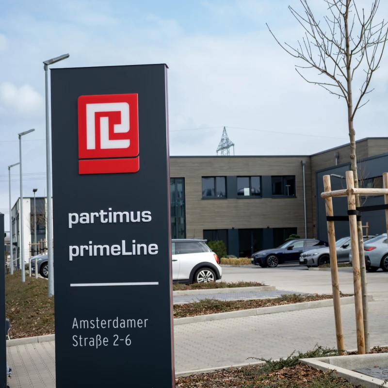 Schwarzes Schild mit weißer Schrift "partimus primeLine" und rot weißem Patimus Logo vor der Einfahrt zum Unternehmenssitz.